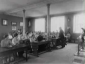 Interiör från A-salen på Katrinebergs folkhögskola. De kvinnliga eleverna sitter vid skolbänkar och rektor Ludvig de Vylder står lutad vid en av de främre. Det är troligen en klass från lanthushållsskolan. 1910-talet.