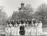 Prästen Johansson (i mitten) med konfirmander. Kyrkan i bakgrunden, delvis dold bakom träd. Sittande från vänster: ?, Stina Andersson, Karin Andersson, övriga okända. 2:a raden: Dagny Andersson från Fagerhult, Ingrid Andersson från Fagerhult, Anna-Lisa Johansson från Ryareås, Iris Andersson från Böstekullen, Karin Johansson från Djupdalen, ?, ?.