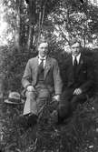 Två män sitter i en en skogsbacke och håller varandra i handen.