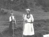 En kvinna och en flicka fotograferade iklädda folkdräkter? stående i en skogsbacke.