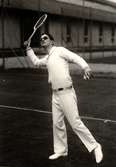 Nordiska Kompaniet visar tenniskläder oktober 1928. Ung man iförd tenniskläder och solglasögon svingar en tennisracket, i andra handen håller han en boll.