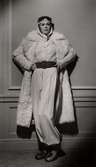 En kvinnlig modell poserar i en vit byxdress med ett brett skärp i midjan. Ovanpå byxdressen bär hon en vit päls. Varuhuset Nordiska Kompaniet i Stockholm visar vintermode 1936.