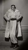 En kvinnlig modell poserar i en vit byxdress med ett brett skärp i midjan. Ovanpå byxdressen bär hon en vit päls. Varuhuset Nordiska Kompaniet i Stockholm visar vintermode 1936.