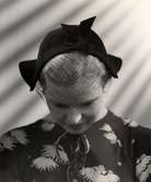 Hattmode visas på varuhuset  Nordiska Kompaniet i Stockholm 1936.
En ung kvinna poserar med nedböjt huvud iförd en liten hatt med uppvikta brätten, liten rosett mitt fram och stormaskigt flor framför ansiktet. Hon bär en storblommig klänning med knytband i halsen.