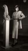 Dammode visas på varuhuset Nordiska Kompaniet i Stockholm 1937. En kvinnlig modell poserar iklädd dräkt brevid en skulptur i föreställande av två fiskar. Kjolen är vadlång, blusen har knytband, jackan är av ledig modell. Till detta bär hon herrhatt, handskar och pumps med klack.