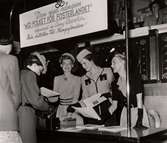Kungafonden på Nordiska Kompaniet 1943. Tre leende kvinnor står bakom ett bord, under en skylt med texten 