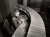 Grammofonavdelningen vid varuhuset Nordiska Kompaniet i Stockholm 1947. En ung kvinna i tweeddräkt sitter vid ett svängt mixerbord av trä. Framför henne flera stycken skivspelare, knappar och reglage. Hon trycker på en knapp.