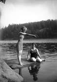 Två kvinnor i baddräkt på sten vid vattnet. Den ena gör sig beredd att dyka i.