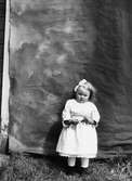 En liten flicka i lång vit klänning poserar framför en fondvägg. Lima, Dalarna