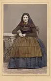 En kvinna i folkdräkt poserar sittande iklädd sjalett och randigt förkläde. Genarp, Skåne. Kolorerat i efterhand. Nordiska museet iv.nr. 18536C.