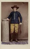 En man poserar stående iklädd en folkdräkt. Han vilar armen på en kolonn, i andra handen håller han en käpp. Fotografiet kolorerat i efterhand.