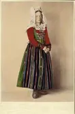 Kvinna i helfigur klädd i högtidsdräkt för gift kvinna från Vemmenhögs härad, Skåne. Handkolorerat fotografi