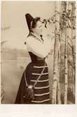 Porträtt av ung kvinna i folkdräkt från Dalarna som står och ristar i en trädstam.