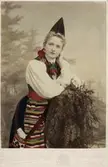 Kvinna klädd i folkdräkt från Dalarna står och lutar sig mot en stubbe. Handkolorerat ateljéporträtt.