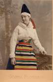En kvinna poserar i en folkdräkt från Dalarna, med en kort vinterjacka till. Handkolorerat fotografi.