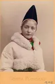 Bröstbild av en ung kvinna i folkdräkt från Rättvik i Dalarna, med en pälsbesatt vit jacka. Kolorerat fotografi.