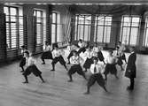 Gymnastik. Flickor i vita blusar och vida svarta knäbyxor gymnastiserar under överinseende av en manlig lärare. Nya Flickskolan, Sala