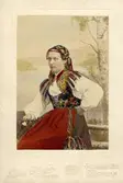 En ung kvinna poserar i en folkdräkt med schalen instoppad i västen. Handkolorerat fotografi.