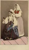 Dräktdockor med huvuden skulpterade av C A Söderman, visade på världsutställningen i Paris 1867. Två kvinnor i folkdräkter från Blekinge, varav en hjälper den andra med huvudbonaden. Handkolorerad version.