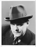 Man med hatt. Porträtt av medelålders man iklädd kritstrecksrandig kostym, mönstrad slips och hatt, brättet skuggar hans ansikte.