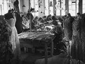Interiör från Franska damskrädderiet på Nordiska Kompaniet. Sömmerskor i arbete. Vid arbetsbord sitter en grupp kvinnor och syr för hand. I fonden stora höga fönster där ljuset silar in, ovanför borden hänger många lampor. I förgrunden står provdockor, så kallade mannekänger, där plaggen provas ut. 7 maj 1947.