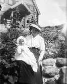 Kvinna sitter på stenmur med spädbarn i famnen. Fiskebäckskil, Bohuslän.