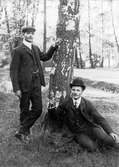 Två unga män i utomhusmiljö, den ene sittande med promenadkäpp och plommonstop, den andre med keps lutandes mot en björk.