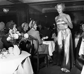 Konfektionsvisning i lunchrummet på Nordiska Kompaniet.
Modell i aftonklänning med paljettbroderi passerar åskådarna. Publiken sitter vid uppdukade bord med vita dukar, NK-servis och blommor i vas. De flesta kvinnorna bär hatt.