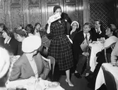 Sahlénsvisning i lunchrummet på Nordiska Kompaniet.
Modell klädd i rutig klänning med skärp och knappar, hatt och ljusa pumps passerar åskådarna. Modellen håller en skylt som visar 116SEK. Publiken sitter vid bord och tittar på modellen.