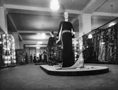 Interiörbild från Nordiska Kompaniet. Skyltdockor i hellånga klänningar på podie i mitten och klänningar på galgar längs sidorna.