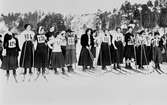 Startfältet i damernas skidtävling i Saltsjöbaden 1910. Damernas tävling, Djurgården IFK Djursholm. Tvillingsystrarna Tora och Sigrid Wiking (f. 1893) från Djursholm deltog. Sigrid har nr 126 och står i främre ledet, Tora har en likadan tröja och står i bakre raden. Systrarna hade redan vintern 1909 som pionjärer börjat använda byxor vid skidåkning.