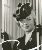 Porträtt av kvinna klädd i hatt med veck, kavaj, blus, handskar och brosch.