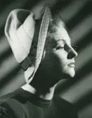 Porträtt av kvinna i hatt i svagt rosa filt med stickningar på kullen, garnerad med marinblått band av Claude St Cyr.