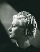 Porträtt av kvinna med hatt i smultronfärgad filt med brätte av flätade sidenband i en mängd färger. Garneringen bildas genom att banden avslutas i nedhängande öglor. Modell efter Simone Cange.