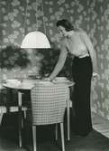 Helporträtt av kvinna i hellång klänning med skärp, står vid dukat bord.