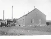 Bebyggelsevy, industrifastighet. En större tegelbyggnad och kompletteringsbyggnader/uthus med två skorstenar.