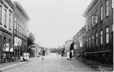 Stensatt stadsgata i Varberg. Stadsbebyggelse. Hus med träfasader och tegelhus. Södergatan mot öster. Husen till höger närmast kameran ligger troligen i kvarteret Bokbindaren. T v Oskar Johanssons Bleck & Plåtslageri. Några fotgängare. Bild utan år.