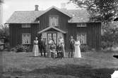 Familjebild med åtta personer framför mangårdsbyggnaden med sticktak (pärt), , två skorstenar och glasveranda med snickarglädje.