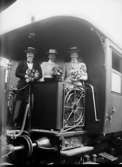 Tre leende kvinnor med blomsterbuketter i händerna står på en plattform bakpå en järnvägsvagn