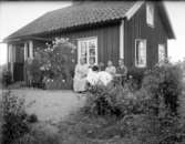 Artur och hans mor Anna Pettersson sitter tillsammans med en annan kvinna och hennes son (?) vid ett dukat kaffebord utanför ett bostadshus. 
I förgrunden breder den prunkande trädgården ut sig.