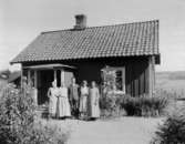 Fyra kvinnor, bl. a. Anna Pettersson och Augusta Forsberg, och en man, Artur Pettersson, poserar utanför en stuga. I förgrunden ses delar av en trädgård med ett stort antal olika växter.