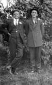 Två herrar i kostym och hatt stående framför ett buskage