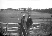 En man och en kvinna vid en gärdesgård. En gård med omgivande fält syns i bakgrunden.