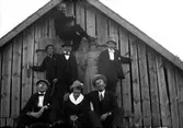 Åtta personer framför en husgavel av trä. Fotografens anmärkning: Gruppbild mossen.