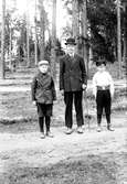 En man tillsammans med två pojkar, i bakgrunden barrskog samt ett framskymtande trähus.
