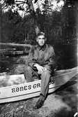 En ung man i soldatuniform sitter i en halvt uppdragen båt med texten ROBES GA .. OS. I bakgrunden syns barrskog samt en roddbåt vid sjökanten. Lungkliniken, Eksjö.
Fotografens anmärkning 