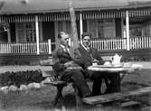 Två män sitter på en bänk vid ett bord med kaffekanna och koppar. I bakgrunden ett trähus med en stor veranda.