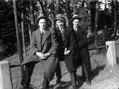 Tre män i rock och hatt, sitter på ett metallräcke. I bakgrunden barrskog.