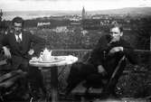 Två män sitter vid en uteservering med utsikt över Jönköping. Bordet är dukat med koppar och fat, på en bricka står bland annat en kaffekanna.