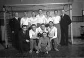 Tolv män, nio idrottsklädda och tre i kostym, Hallby handbollslag, står uppställda i ett handbollsmål.
Översta raden: Johansson (4), Rundgren (5), Ordförare Moberg (7).
Nedre raden: Gustavsson (2), Adolf Johansson (Fiskarn) (3), Eilert Lind (längst till höger).